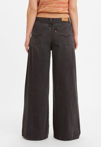 Person wearing high-waist wide-leg black jeans with back pockets and an orange knit sleeveless top, shown from behind.