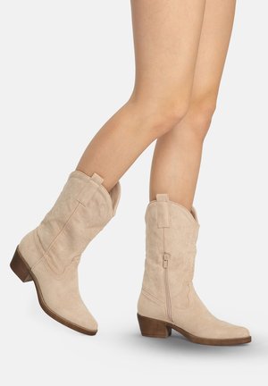 Beine, die beige Wildleder-Cowboystiefel bis zur Mitte der Wade mit seitlichen Reißverschlüssen und niedrigen braunen Absätzen tragen, vor einem schlichten hellen Hintergrund posieren.