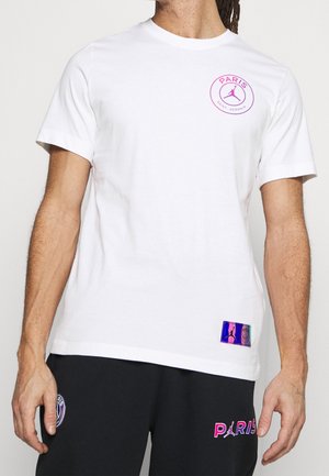 Mann trägt weißes Paris Saint-Germain T-Shirt mit pinkem Logo auf der Brust und schwarze Shorts mit pinkem "Paris" Schriftzug und Logo.