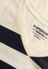 Koszulka w kolorze ecru z granatowymi paskami, dekolt w serek, z logo "Superdry Premium" i napisem "Finest Manufactured Goods New York, N.Y." wewnątrz kołnierzyka.