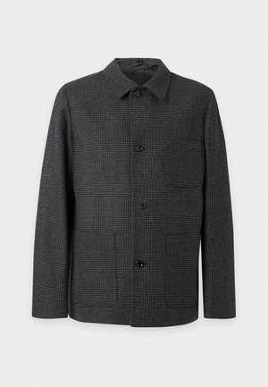 Jeune homme portant une veste à carreaux foncés boutonnée, un pantalon noir et des mocassins noirs, debout devant un fond blanc uni.