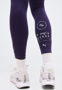 Άτομο που φοράει σκούρο μωβ κολάν με το κείμενο και τα λογότυπα "MY SPORT IS HYROX", λευκές κάλτσες και λευκά παπούτσια Puma για τρέξιμο με σχεδιασμένες φτέρνες.