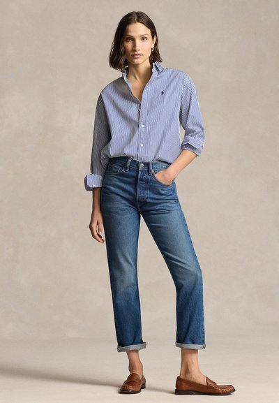 Mujer de pie con la mano en el bolsillo, vistiendo vaqueros azules remangados, camisa de botones a rayas azul y blanca y mocasines marrones.