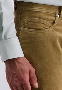 Khakifarbene Cordhosen mit vertikalen Rippen, Seitentaschen und einem silbernen Knopfakzent, kombiniert mit einem weißen Hemdärmeln mit Punkten.