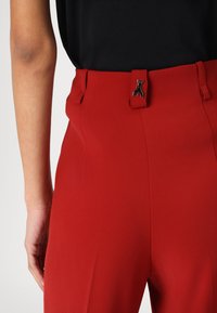 Crvene krojene hlače s glatkom teksturom, s crnim metalnim kopčanjem na struku i elegantnim, prilagođenim dizajnom.