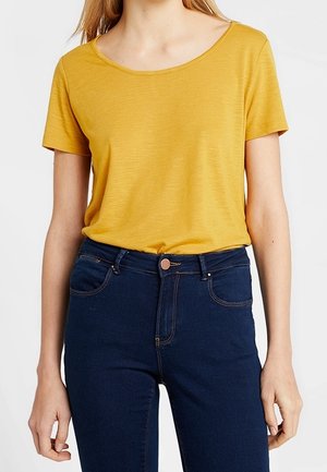Musztardowożółta koszulka z krótkim rękawem i okrągłym dekoltem, połączona z ciemnoniebieskimi dżinsami z guzikami i przednimi kieszeniami.