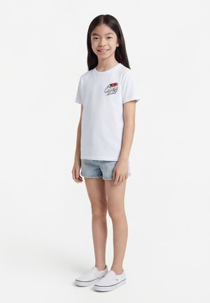 Jong meisje dat staat, gekleed in een wit T-shirt met korte mouwen met een kleine rood-zwarte afbeelding, lichte spijkershorts en witte instapschoenen.