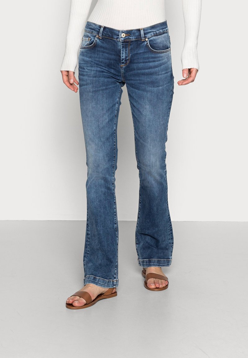 Personne portant un jean bootcut bleu, un haut blanc côtelé à manches longues et des sandales plates marron, debout sur un sol uni.