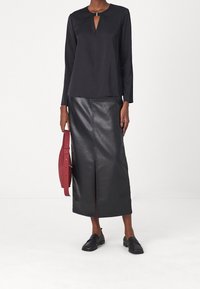 Haut noir à manches longues avec un détail œillet, associé à une jupe en cuir noire taille haute avec une fente à l'avant. Tenant un sac à main rouge.