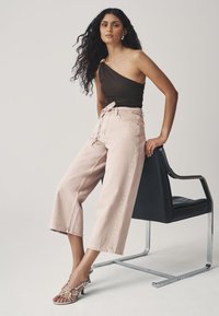 Barna egyrészes vállpántos felső, világos rózsaszín bőszárú farmernadrág, ezüst szandál pántokkal, fekete széken ülve, minimalista háttérrel.