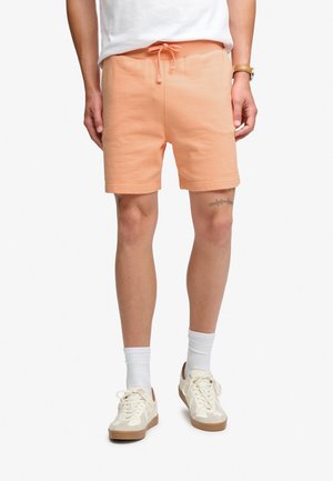 Homme portant un short orange clair, des chaussettes blanches classiques, des baskets blanches et un t-shirt blanc, debout, les mains détendues le long du corps.