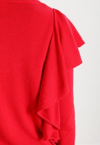 Tissu rouge vif avec détail de volants superposés sur l'épaule d'un vêtement en tricot, sur un fond clair uni.