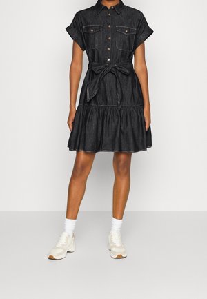 Femme vêtue d'une robe en jean noire à manches courtes avec poches avant et ceinture à la taille, associée à des baskets blanches et des chaussettes, debout sur un fond uni.