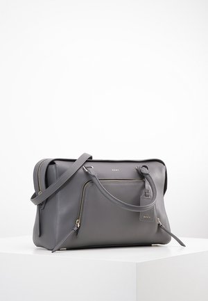 Sac à main en cuir gris DKNY avec poche zippée à l'avant, double poignée et bandoulière réglable sur surface blanche avec fond blanc.