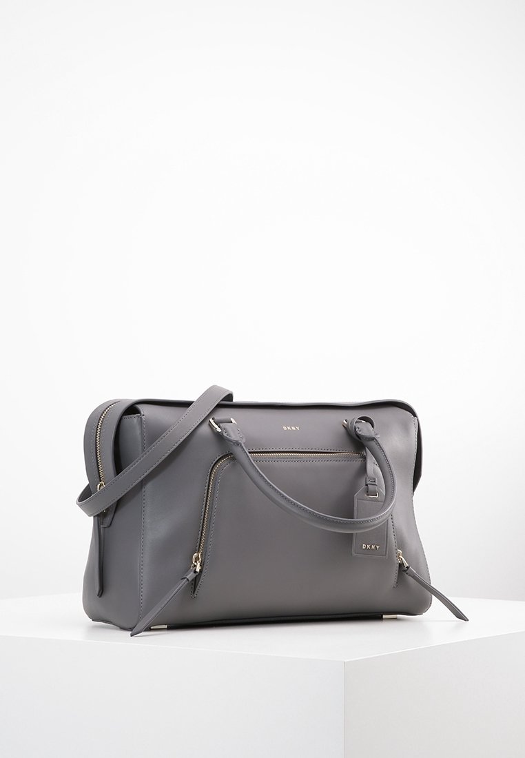 Sac à main en cuir gris DKNY avec poche zippée à l'avant, double poignée et bandoulière réglable sur surface blanche avec fond blanc.