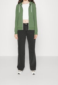 Osoba v zelené zipové mikině, bílem krátkém topu, volných černých džínách a bílých teniskách stojící proti jednobarevnému světlému pozadí.