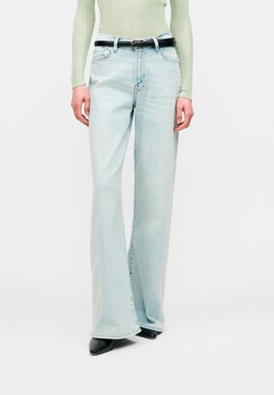 Άτομο που φορά ανοιχτό μπλε ψηλόμεσα τζιν καμπάνα, μαύρη ζώνη, μαύρα μυτερά παπούτσια και ανοιχτό πράσινο ριμπ πλεκτό μακρυμάνικο τοπ.