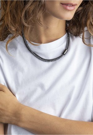 Donna vestita con una camicia bianca e una collana di perline nera, con il braccio incrociato, che mostra parte del suo viso e del collo.