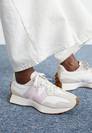 Weiße und beige Sneaker mit pinken Akzenten, getragen zu weißen Hosen mit weitem Bein auf blauem Teppichboden.
