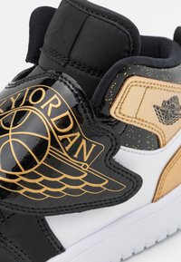 Černé lakované tenisky s zlatými akcenty, bílou mezipodešví a embosovaným logem. Jsou viditelné texturované černé a béžové prvky.