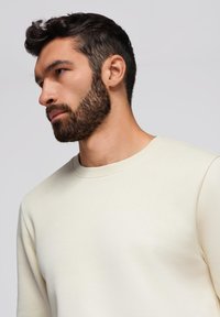 Cremefarvet sweatshirt med rund hals, blød tekstur og tilpasset design, vist iført af en mand med skæg og kort hår.