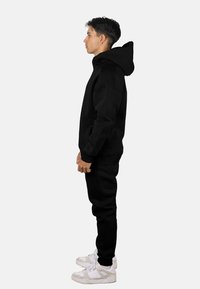 Sudadera con capucha negra con bolsillo frontal, combinada con pantalones jogger negros a juego. Presenta una capucha grande y puños acanalados. Combinado con zapatillas blancas.