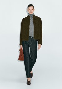 Žena v tmavě zelené zapínací bundě, šedém roláku, černých džínách, hnědých botách a s hnědou kabelkou na neutrálním pozadí.