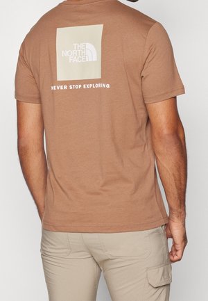 Man draagt een lichtbruine The North Face t-shirt met logo en de tekst ‘Never Stop Exploring’ op de rug, gecombineerd met beige cargobroek.