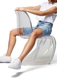 Persona che indossa sneaker bianche, pantaloni corti in denim e una t-shirt bianca, seduta su una sedia moderna in plastica trasparente, mentre tiene il poggiatesta.