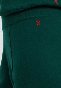 Donkergroene stof met een zachte textuur en een rode geborduurde "X"-detail. Zichtbare naden en een gevouwen rand aan de bovenkant.