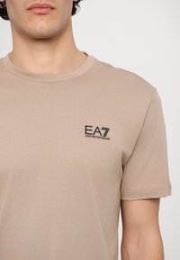Beige tričko s kulatým výstřihem vyrobené z elastické bavlny, s malým černým logem EA7 na levé části hrudi. Krátké rukávy, hladká textura.