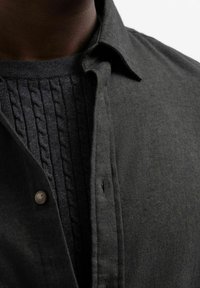 Camisa de botones de color gris oscuro, colocada sobre un suéter oscuro texturizado con un patrón de punto de cable. Cuello y botones visibles en la zona del escote.