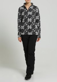 Fleece-Pullover im schwarz-weißen geometrischen Muster, hoher Kragen und Reißverschluss. Kombiniert mit schwarzen Hosen und schwarzem Schuhwerk. Glatte Textur.