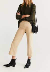 Camisa de botón verde oscuro, pantalones cortos beige y botas negras hasta el tobillo. Pequeño bolso cruzado negro con detalle de hardware circular.