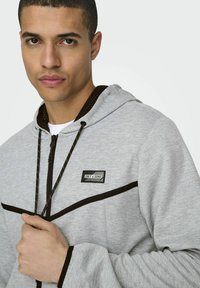 Sudadera con cremallera en gris, con acentos en negro y un parche de logo en la parte frontal, con capucha ajustable y textura suave.