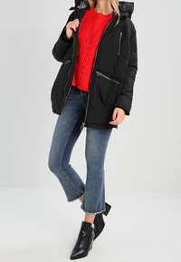 Veste noire rembourrée avec capuche, fermeture éclair, garnitures dorées, poches latérales, portée sur un pull en tricot rouge et un jean évasé bleu. Bottes noires à la cheville.