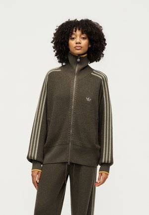 Donna con capelli ricci che indossa un maglione in maglia verde oliva scuro con zip, con tre strisce beige sulle maniche e pantaloni in maglia abbinati.
