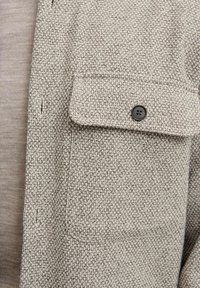 Vævet grå skjorte med en tekstureret finish, der har en brystlomme og en brun knap. Underneath er et lys beige lag synligt.
