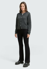 Grau gestrickte Strickjacke mit Rundhalsausschnitt, vorne mit Knöpfen, taillierte Passform, kombiniert mit schwarzen Schlaghosen und schwarzen Plateauschuhen.