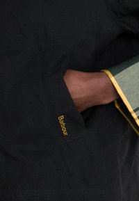 Casaco preto com um bolso lateral que apresenta bordado amarelo "Barbour". O interior tem um forro com riscas verdes e amarelas visíveis na manga.