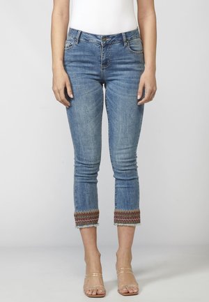 Vrouw draagt lichtblauwe cropped jeans met meerkleurige franjes aan de zoom en doorzichtige sandalen met bandjes, tegen een eenvoudige achtergrond.