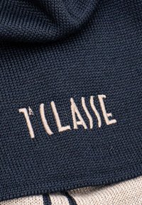 Indumento lavorato a maglia in blu navy con ricami beige più chiari che riportano la scritta "1ère CLASSE." Mostra un tessuto strutturato con punti e pieghe visibili.