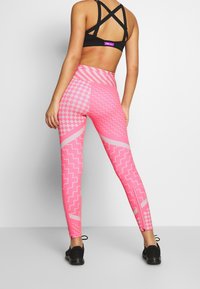 Różowe legginsy w wzory z mieszanką motywów zygzaków i kratek, wykonane z elastycznego materiału, połączone z czarnym stanikiem sportowym i czarnymi butami.