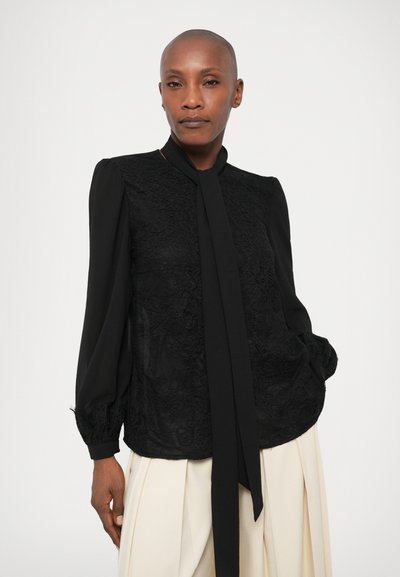 Melnā blūze ar strukturētu rakstu, garām piedurknēm un sasietu apkakles detaļu. Apvienota ar gaišām biksēm. Gluda auduma un brīvs griezums.