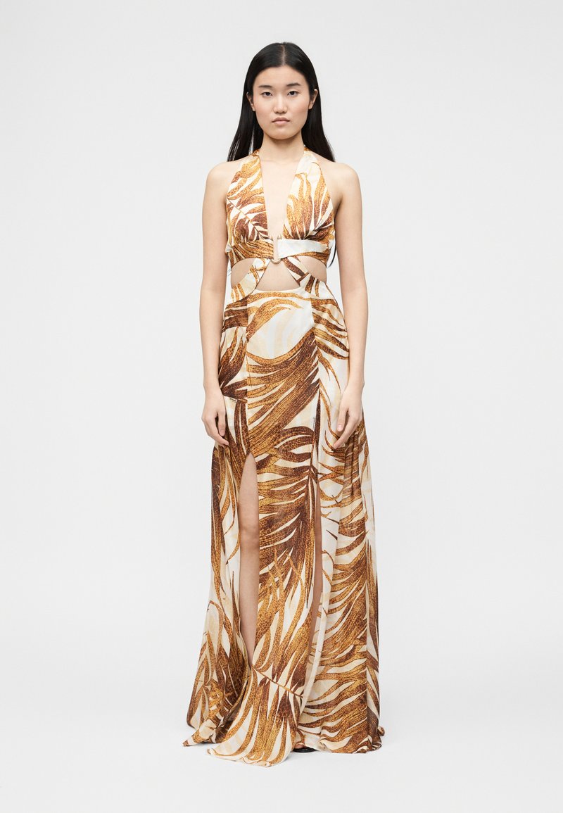 Femme portant une robe maxi sans manches à col licou avec un imprimé feuilles marron, des découpes à l'avant et une fente profonde à l'avant, debout devant un fond uni.