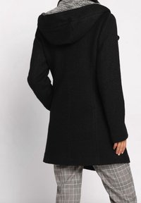 Zwarte jas met een gestructureerde afwerking en een capuchon, met een grijze binnenvoering. Gecombineerd met pantalon in een ruitpatroon.