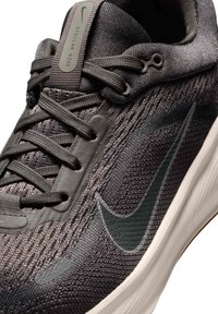 Šedé pletené športové topánky s textúrovaným zvrškom, čiernymi akcentmi, šnurovaním a hrubou bielou medzipodrážkou. Logo Nike viditeľné na boku.