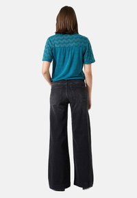 Türkiser Oberteil mit strukturiertem Spitzeneinsatz hinten, kombiniert mit hochtaillierten, weit geschnittenen schwarzen Jeans. Glatter Stoff, lockerer Schnitt. Lange Ärmel.