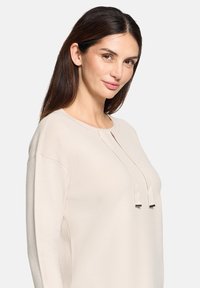 Jasno beżowa bluzka z długim rękawem i okrągłym dekoltem, z dwoma dekoracyjnymi sznurkami z przodu oraz gładką, miękką fakturą.