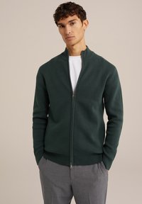 Tumši zaļš nepiepildāms žakets ar augstu apkakli, izgatavots no teksturēta auduma. Apvienots ar gaiši pelēkiem biksēm, ar sānu kabatām.